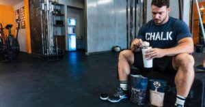 Man preparing a drink in a shaker bottle at the gym, with a bag of nutritional supplement on the floor.
