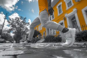 Person in white sneakers doing knee touch exercises on a city sidewalk with selective color highlighting the yellow building and wet ground.