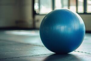 A large blue stability ball sits on a black rubber mat in a room with natural light streaming through large windows.