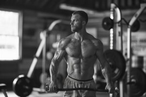 A muscular man holding a barbell in a gym, looking to the side, with weightlifting equipment in the background, debunking gym myths, in black and white.