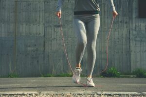 A person in gray sportswear and white sneakers using a jump rope on a concrete surface beside a gray wall.