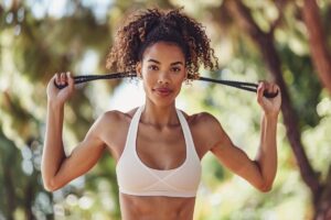 A young woman stretching a resistance band over her head in a park, wearing a white sports bra, with curly hair and a focused expression, feeling her shoulders sore from the workout.