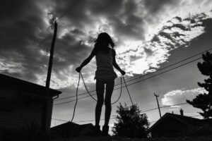 Silhouette of a child engaged in a high-intensity jump rope workout outside, with a cloudy sky and power lines in the background.